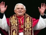 Pope Benedict wearing red, black, and white robes with hands lifted high and smiling