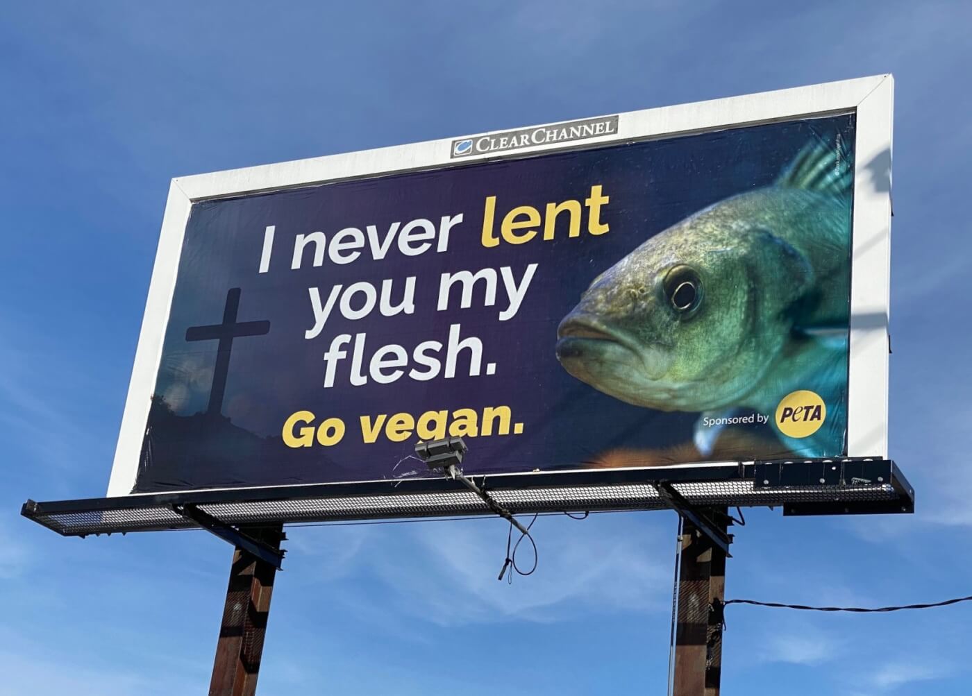 large sign against blue sky with a cross, a fish, and a slogan that says 'I never lent you my flesh. Go vegan.'